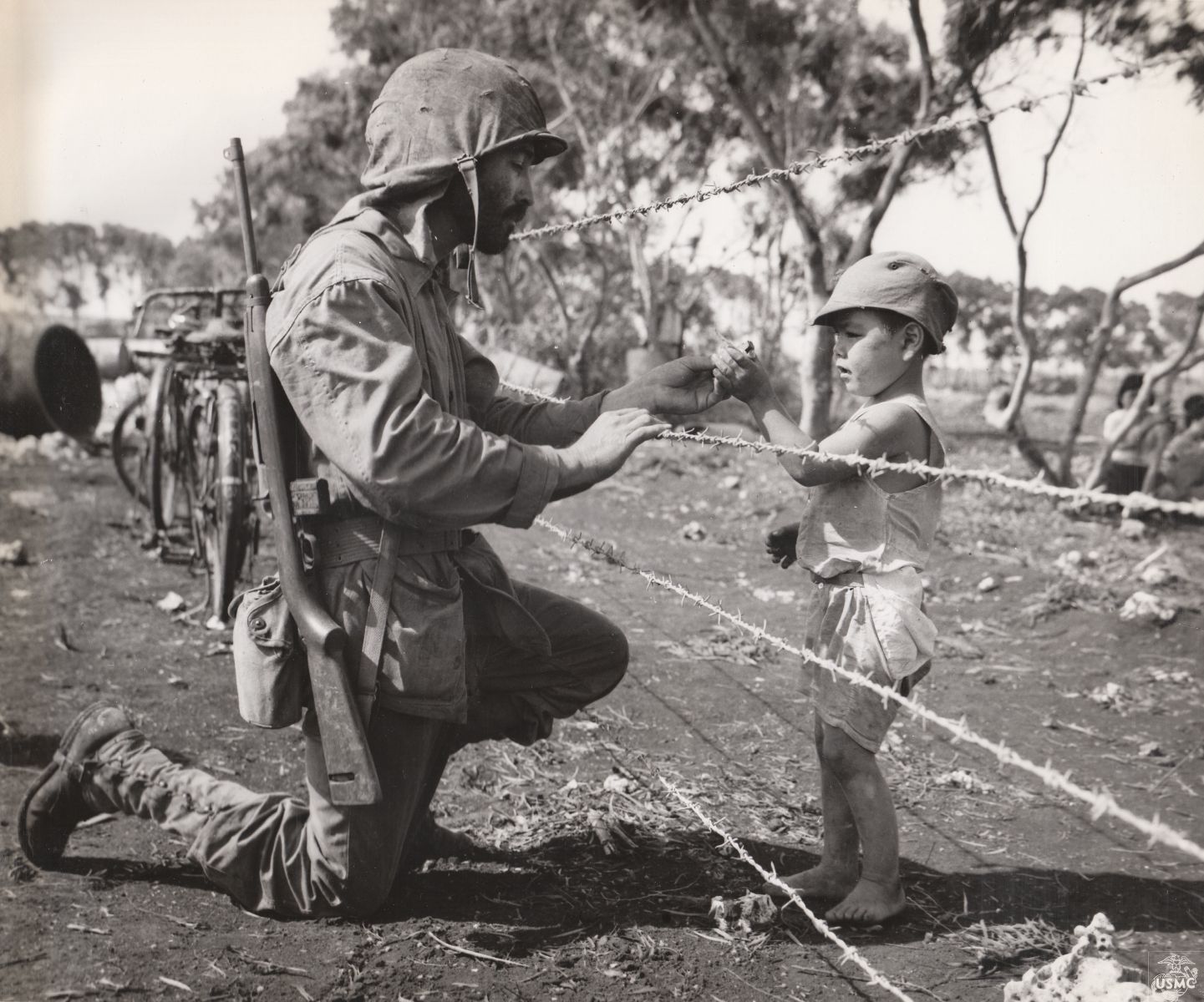 A Soldier in full gear including helmet on his head and a rifle on his backpack kneels down in dirt and reaches through a barbed wire fence with a piece of candy for a young child on the other side who is barefoot with modest clothes and a simple hat on.