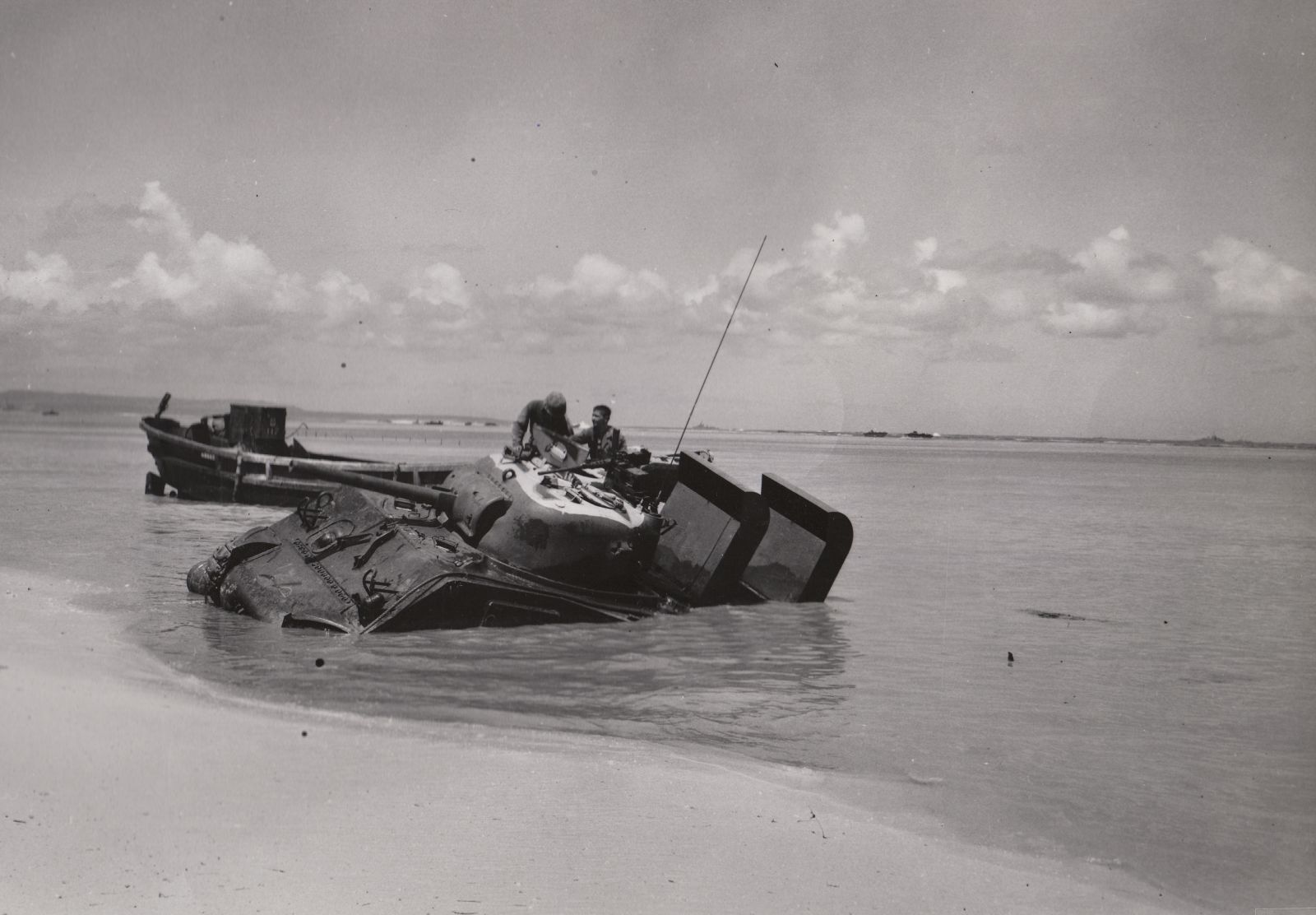 American soldiers climb out of and atop a battle tank that has emerged from the lagoon but sinking into the soft sand of the beach.