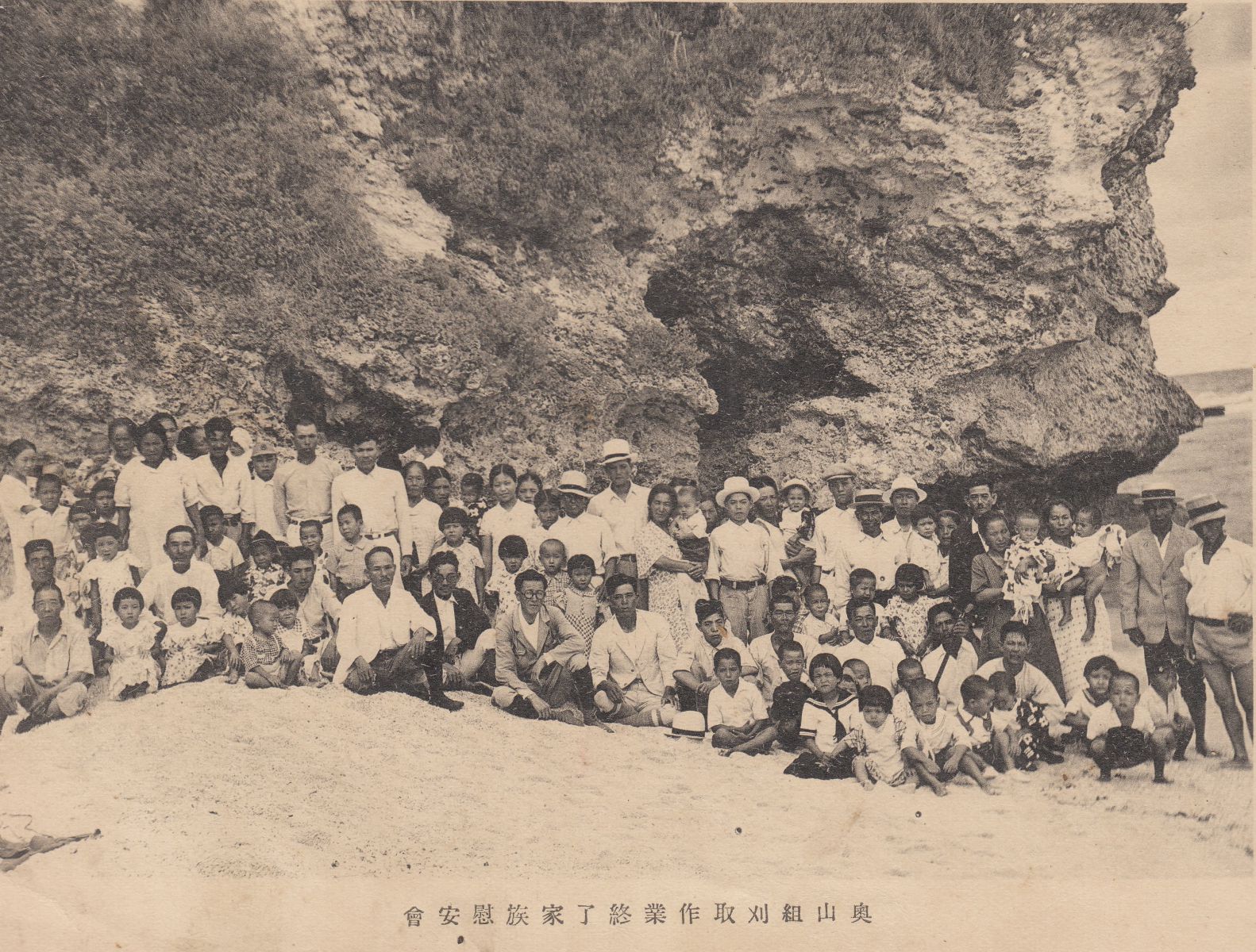 Several families of multiple generations of Japanese people pose for a photo in their holiday clothing standing on a coral beach against the rocky backdrop of a cliff.