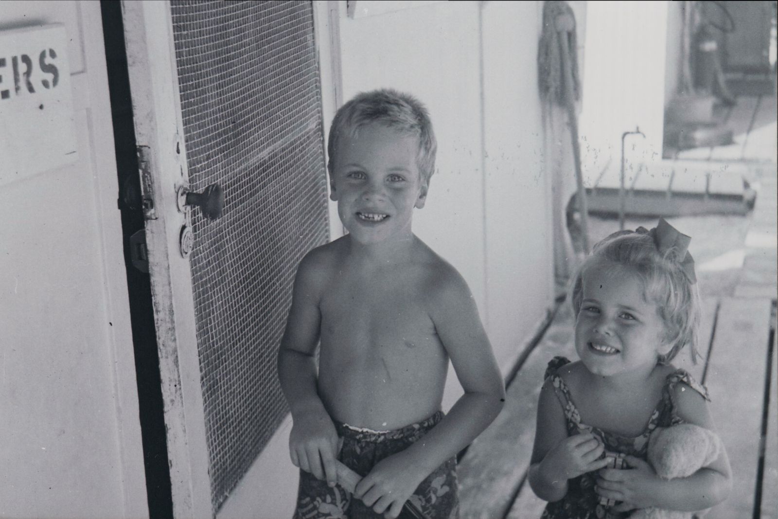 Two little kids with light hair and eyes and fair skin smile next to each other for a photo next to a screen door.