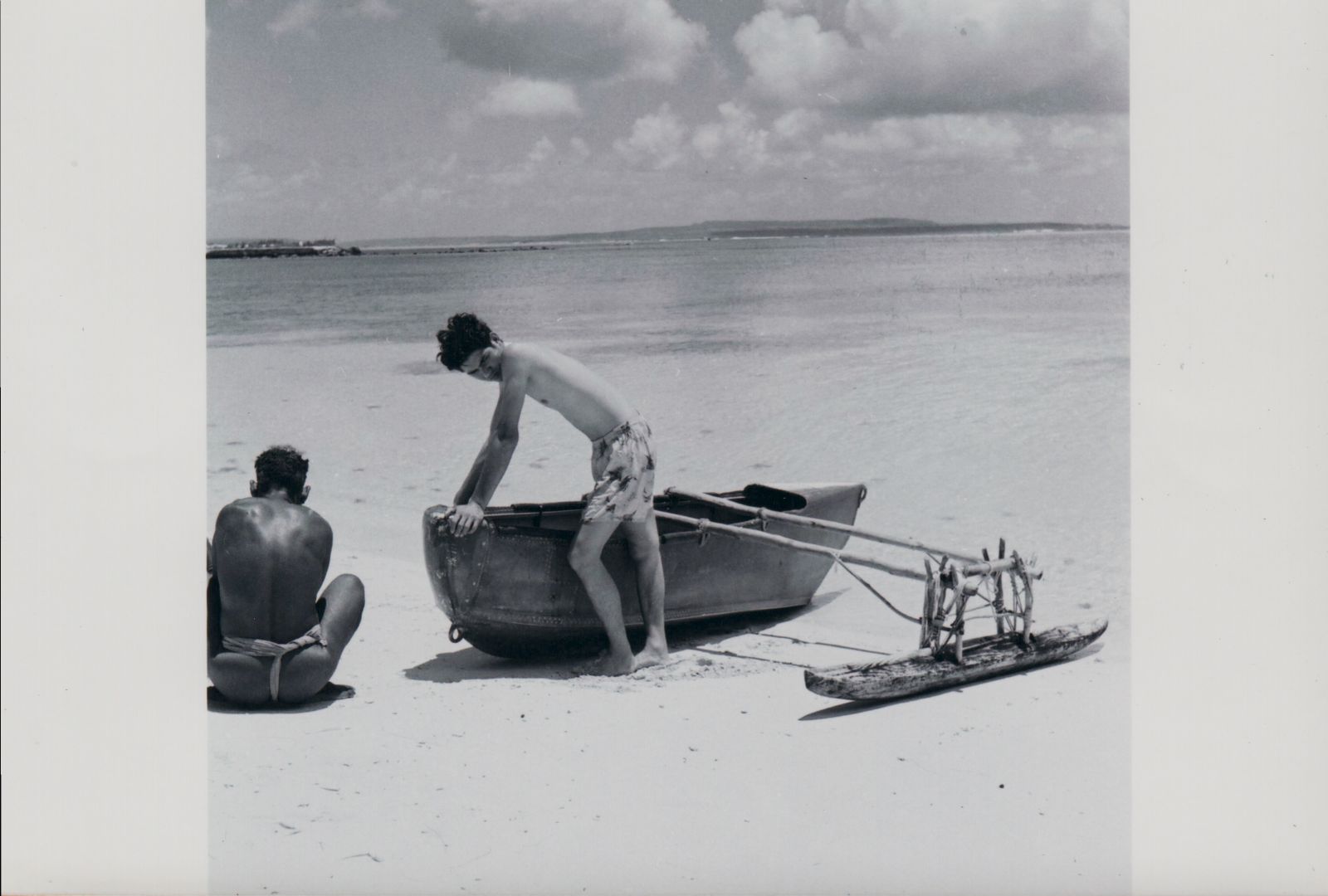 A man in a traditional loincloth sits on the flat, white sand of a beach next to a traditional Chamorro canoe called a galaidi on which another man stands.