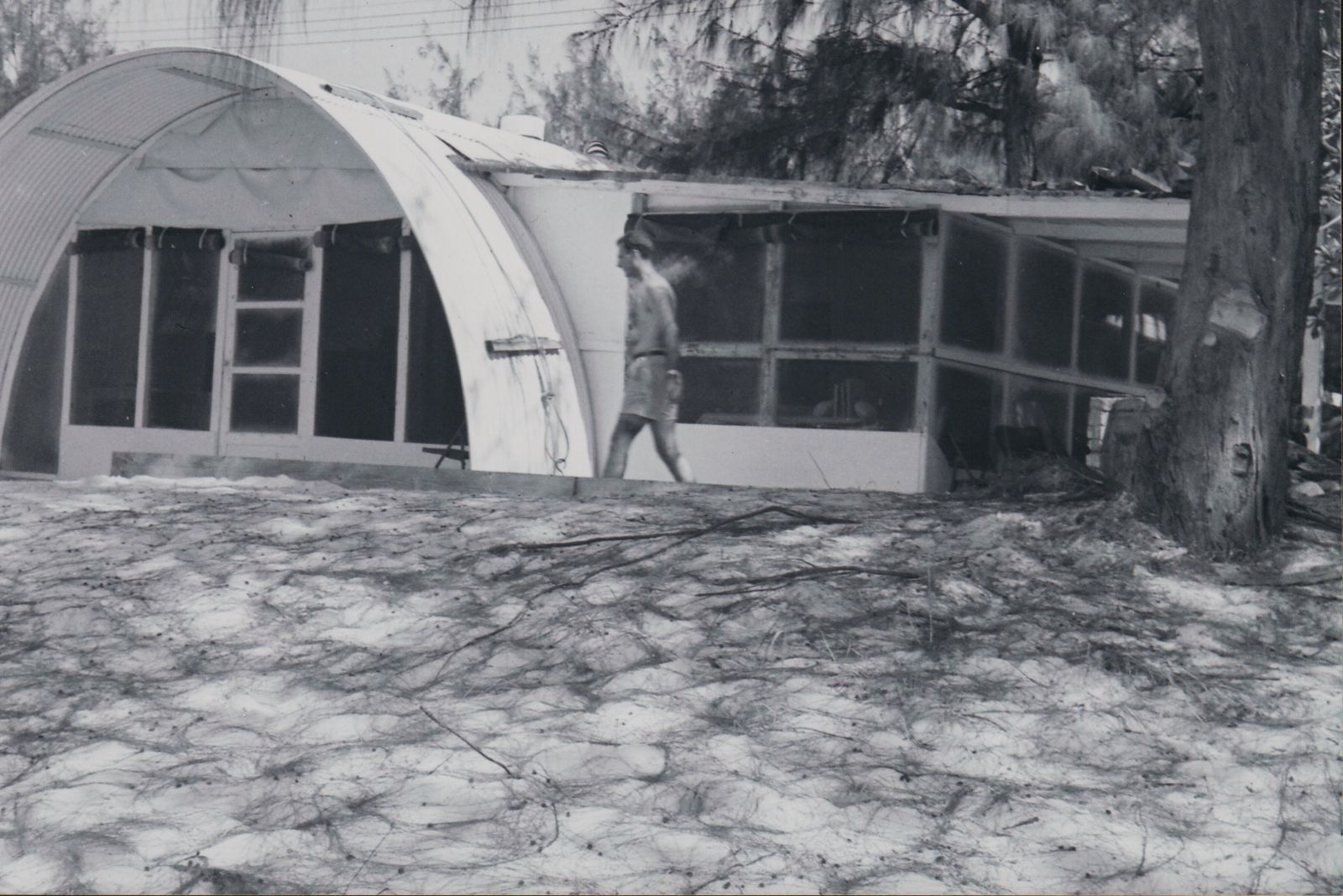 A man crosses the sandy beach yard in front of a quonset hut home with a patio attached.